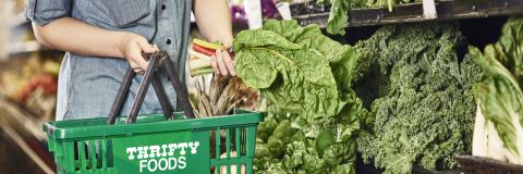 In this image, a lady holds a Thrifty food basket and vegetables in her hands.